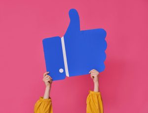 blue thumbs up cut-out being held by a pair of hands on a hot pink background; community engagement; social media likes;