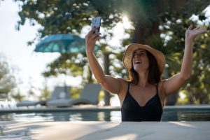 Girl in black hat and swimsuit with cell phone in the pool talking and smiling while making a video call; App concept; Loyalty App